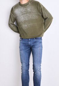 Sudadera color caqui con una textura desvanecida y cuello redondo, combinada con jeans azul ajustados. La sudadera presenta detalles de costura en blanco.