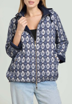 Donna con giacca con zip blu e bianca a fantasia con maniche arrotolate e jeans azzurri.