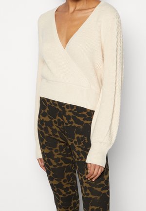 Kvinde iført en cremefarvet V-hals kabelstrikket sweater med lange ærmer og højtaljede sorte og brune bukser med dyreprint