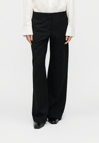 Pantalon noir à jambes larges en tissu lisse, avec une coupe ajustée, des plis sur le devant et des ourlets finis. Porté avec une chemise blanche à manches longues.