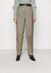 Khaki kalhoty s vysokým pasem a volným střihem, s předními skladbami, černým koženým páskem a jemnou texturou. V kombinaci s černými koženými kotníkovými botami na podpatku.