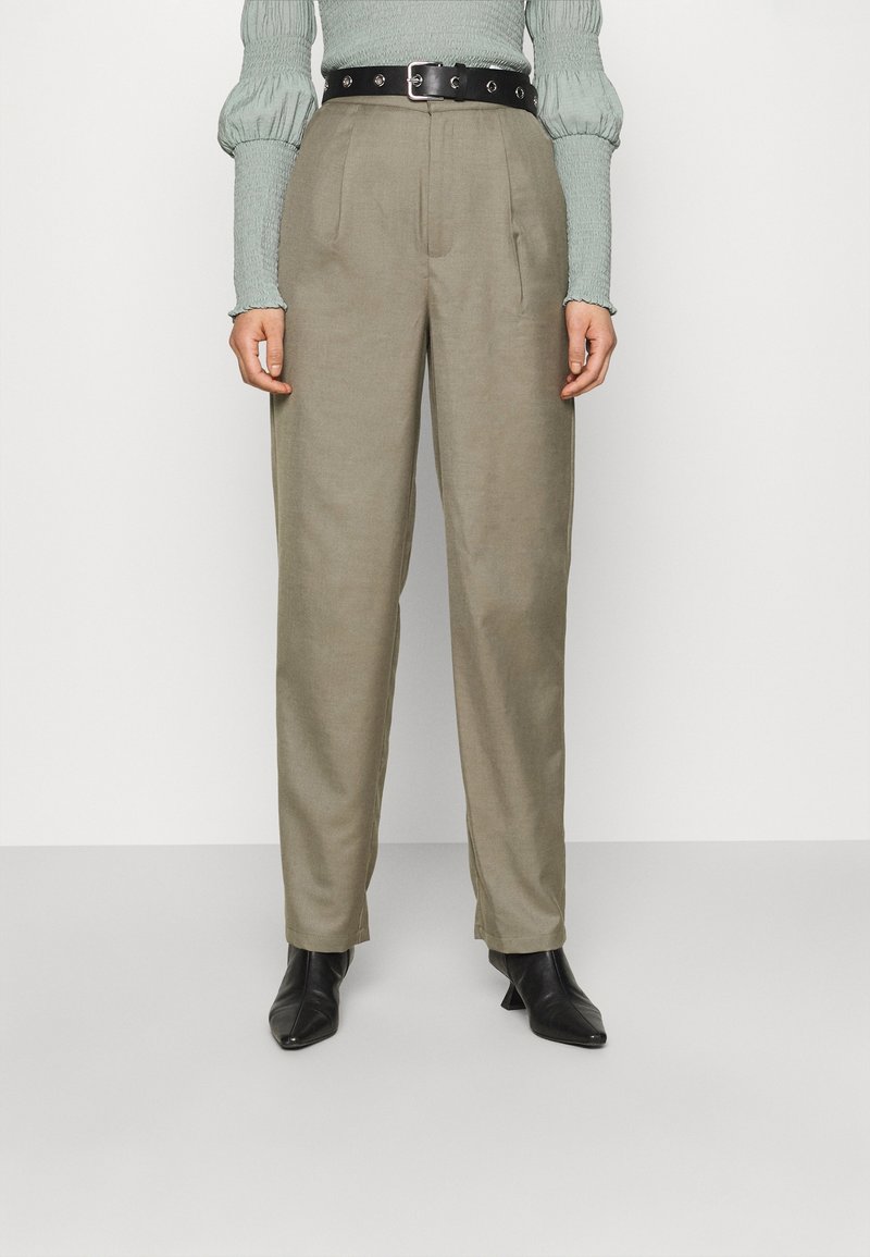 Khaki kalhoty s vysokým pasem a volným střihem, s předními skladbami, černým koženým páskem a jemnou texturou. V kombinaci s černými koženými kotníkovými botami na podpatku.