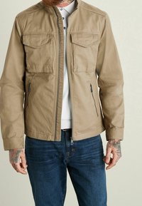 Tan lightweight jacket with a zip front, two chest pockets, side zip pockets, and a stand collar, paired with dark blue jeans.