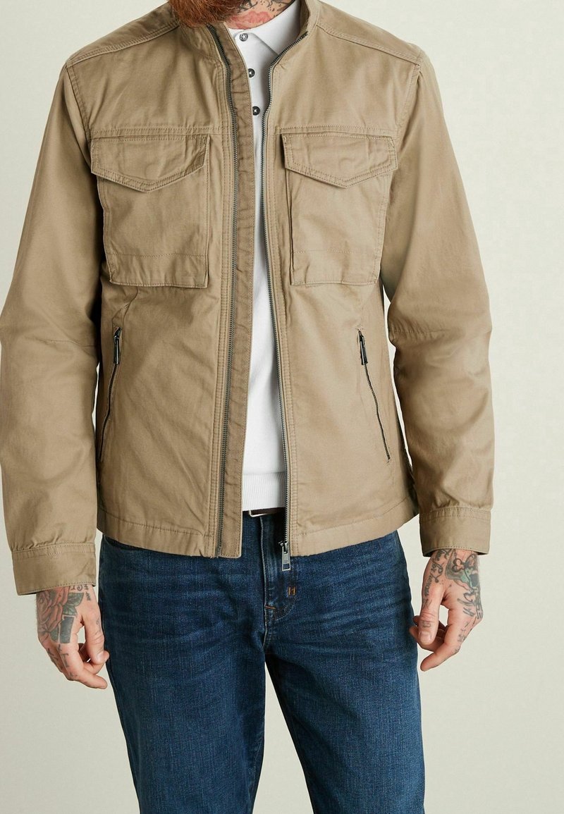 Tan lightweight jacket with a zip front, two chest pockets, side zip pockets, and a stand collar, paired with dark blue jeans.