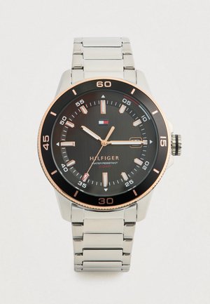 Moška ura na zapestje s srebrnim kovinskim trakom, črnim okroglim številčnicam, oznakami v rožnato zlati barvi, prikazom datuma in logotipom Tommy Hilfiger.