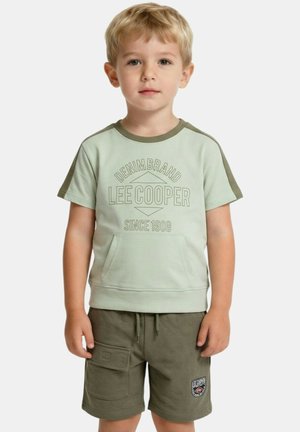 Jeune garçon aux cheveux blonds courts portant un T-shirt Lee Cooper vert clair et un short vert olive, debout devant un fond uni.