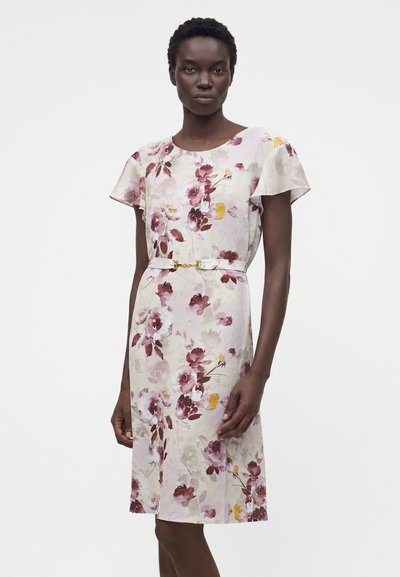 Γυναίκα που φοράει ανοιχτό ροζ φορέμα με λουλούδια μέχρι τα γόνατα, κοντά μανίκια και λεπτή ζώνη στη μέση, στέκεται σε λευκό φόντο.