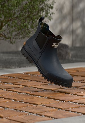 Botte de pluie noire à la cheville, semelle crantée et panneaux latéraux élastiques, dévoilant une doublure tartan et le logo Barbour, posée sur un sol en dalles de bois.