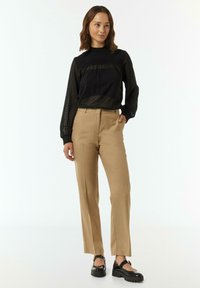 Schwarze Bluse mit langen Ärmeln, strukturiertem Muster und Plissee-Details, kombiniert mit beigen, taillierten Hosen und schwarzen klobigen Schuhen.