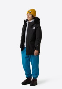 Μαύρο insulated μπουφάν με κουκούλα επενδεδυμένη με γούνα, γκρι φούτερ από κάτω, μπλε παντελόνια τζόγκινγκ και μαύρα ορειβατικά μποτάκια. Κίτρινο ριμπ πλεκτό σκουφάκι, εμφανή λογότυπα.