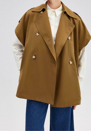 Person trägt einen braunen, doppelt geknöpften, ärmellosen Trenchcoat über einem weißen Hemd mit Knöpfen und blauen Jeans, steht mit den Händen in den Taschen.