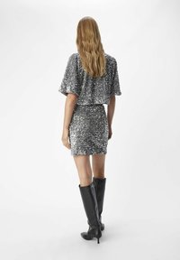 Srebrny błyszczący crop top i pasująca mini spódniczka, w połączeniu z wysokimi czarnymi skórzanymi botkami, widziane z tyłu na białym tle.