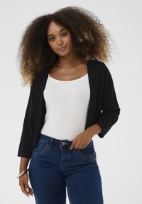 Frau mit lockigem Haar, die ein weißes Tanktop, eine schwarze Strickjacke, blaue Jeans und ein silbernes Armband trägt und vor einem weißen Hintergrund steht.