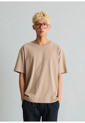 Junge Person mit kurzen blonden Haaren und Brille, trägt ein beiges T-Shirt und dunkle Hose mit Händen in den Taschen vor schlichtem Hintergrund.