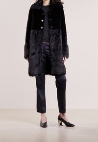 Sort faux fur frakke med en blank finish, der har knaplukning fortil, stribede bukser og sorte hæle med metalliske detaljer.