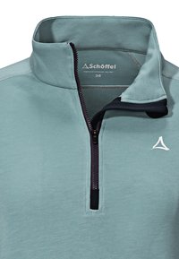 Světle modrý pulovr s čtvrtinovým zipem, který má vysoký límec, černý zip a bílé logo na levé straně, vyrobený z měkké, texturované tkaniny.