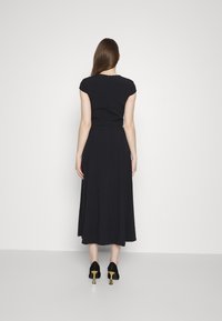 Robe midi noire avec de courtes manches en forme de cap, corsage ajusté et jupe évasée. Portée avec des chaussures noires à talons dorés.