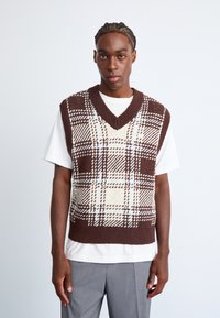 Young man with braided hair wearing a brown and cream plaid knit vest over a white t-shirt and gray trousers against a plain background.