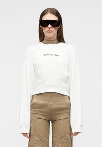 Mujer con grandes gafas de sol negras, sudadera blanca de manga larga y corta, y pantalones de talle alto color beige con cremalleras delanteras.