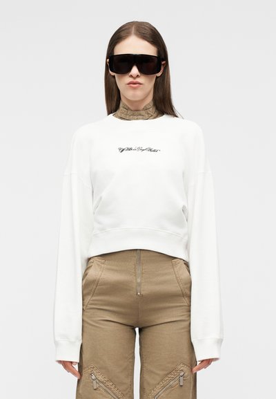 Femme portant de grandes lunettes de soleil noires, un sweat-shirt blanc à manches longues et court, et un pantalon taille haute beige avec des fermetures éclair à l'avant.