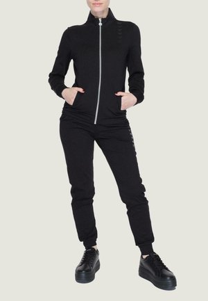 Tuta nera con zip e colletto alto, polsini a costine e tasche laterali. Abbinata a sneakers nere con zeppa. Texture liscia e design aderente.