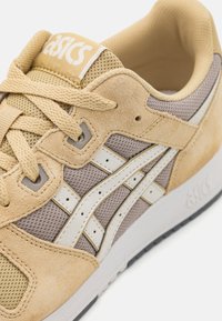 Tamno smeđi i bež tenesici s antilop i mrežastim materijalima; sadrže bijele pruge, okrugle vezice i podstavljeni ovratnik. ASICS logo na jeziku.