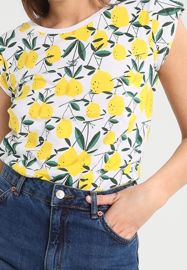 Femme portant une chemise blanche à manches courtes avec un motif de citron jaune et de feuille verte rentrée dans un jean en denim bleu, la main dans la poche.