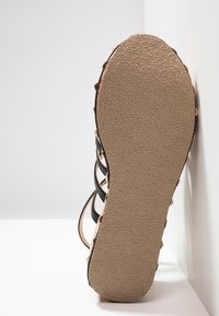 Semelle de sandale plate en caoutchouc beige texturé avec lanières noires, appuyée contre un mur blanc d'angle.