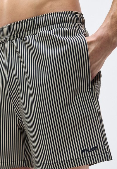 Homme portant un short à rayures verticales noires et beige avec une taille élastique, main dans la poche, torse inférieur et cuisse supérieure visibles.