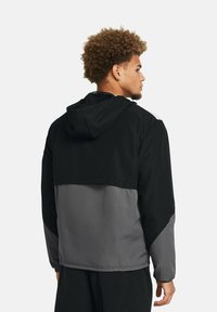 Chaqueta con capucha que presenta una sección superior negra y una sección inferior gris. Hecha de un material ligero con una textura suave y un diseño ajustado.