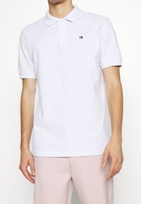 Weißes Poloshirt aus Baumwolle mit klassischem Kragen, kurzen Ärmeln und einem kleinen gestickten Logo auf der linken Brust.