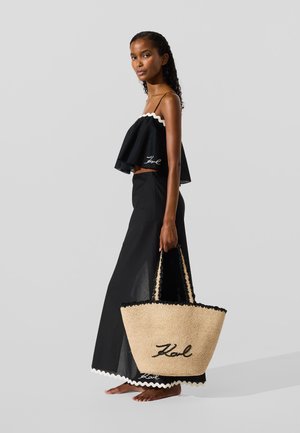 Must olla must crop top lõhkude servadega, mille alla on pandud voolav must seelik, käes on suur õlgkott mustade aksessuaarete ja brodeeritud tekstiga.