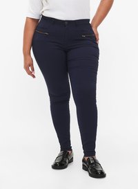 Frau in navy Slim-Fit-Hosen mit goldenen Reißverschlusstaschen, trägt schwarze glänzende Loafers und steht mit einer Hand auf der Hüfte.