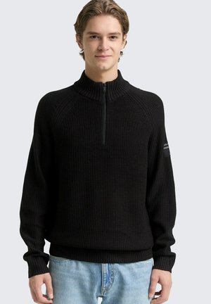 Junger Mann trägt schwarzen gestrickten Pullover mit halbem Reißverschluss und hellblaue Jeans, steht vor einfarbigem hellgrauem Hintergrund.