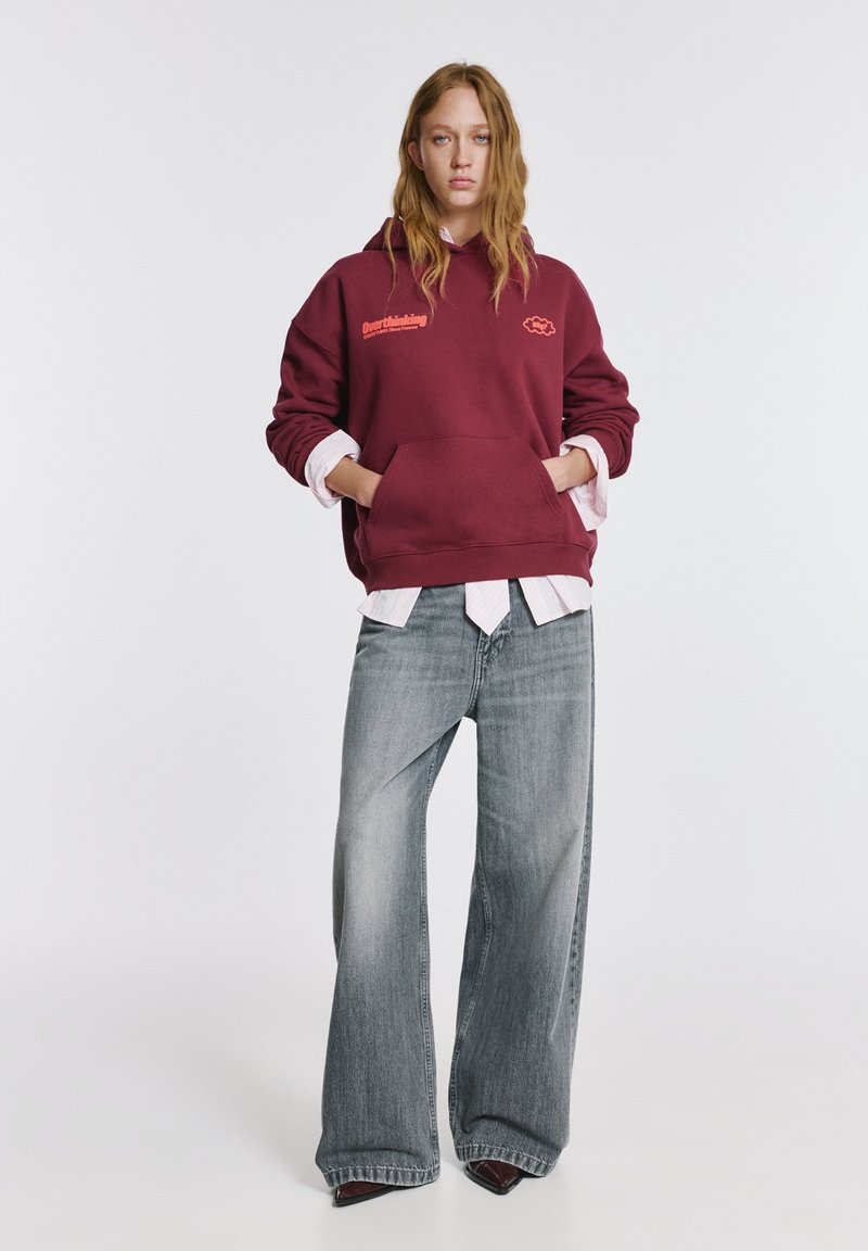 Bordó kapucnis pulóver "Overthinking" felirattal, elülső zsebbel, rózsaszín csíkos ingre rétegezve; széles szárú, kifakult farmernadrággal párosítva.