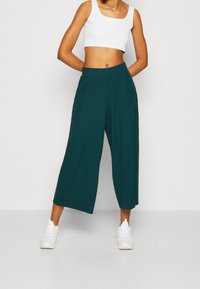 Pantalons larges teal avec une taille élastique, fabriqués en tissu léger. Associés à un crop top ajusté blanc et des baskets blanches.