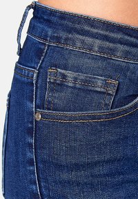 Jeans de mezclilla azul oscuro con un bolsillo frontal, costuras doradas en contraste y una textura suave. Detalle de remache cerca del bolsillo.