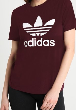 Femme portant un t-shirt Adidas bordeaux avec un grand logo trefoil blanc et du texte, associé à un pantalon noir, sur un fond uni.