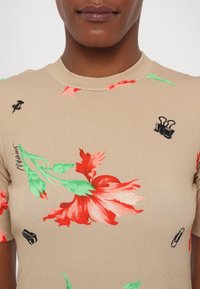 Camiseta beige de manga corta con tejido acanalado, decorada con un patrón floral en rojo y verde, y gráficos distintivos en negro de lápiz, clip y imperdible.