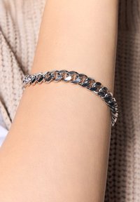 Bransoletka ze srebrnego łańcuszka z owalnymi ogniwami, polerowanym wykończeniem i lekko elastycznym designem, noszona na ramieniu na neutralnym tle.