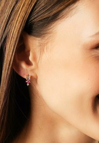 Ohr einer Frau mit goldener Ohrstecker, der drei mehrfarbige Edelsteine – klar, pink und weiß – in vertikaler Anordnung enthält.