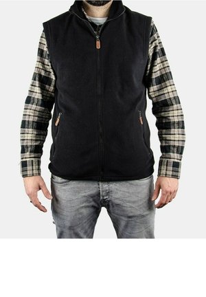 Man draagt een zwart fleecevest met rits over een zwart-beige geruit overhemd en grijze spijkerbroek, staand tegen een effen witte achtergrond.