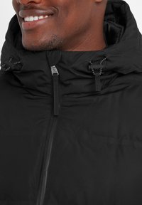 Black insulated jacket with a high collar, drawstring hood, and zipper closure. Textured fabric with visible stitching and hardware accents.