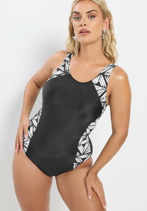 Femme portant un maillot de bain une pièce noir avec des panneaux à motif de feuilles blanches, debout devant un fond clair uni.