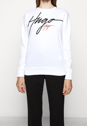 Sudadera blanca con diseño de texto "Hugo" en negro y rojo. Presenta un escote redondo, mangas largas y puños acanalados. Textura de tejido suave.