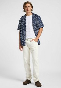 Camicia blu navy a maniche corte con motivo geometrico bianco, indossata sopra una t-shirt bianca, abbinata a pantaloni color crema e zoccoli marroni.