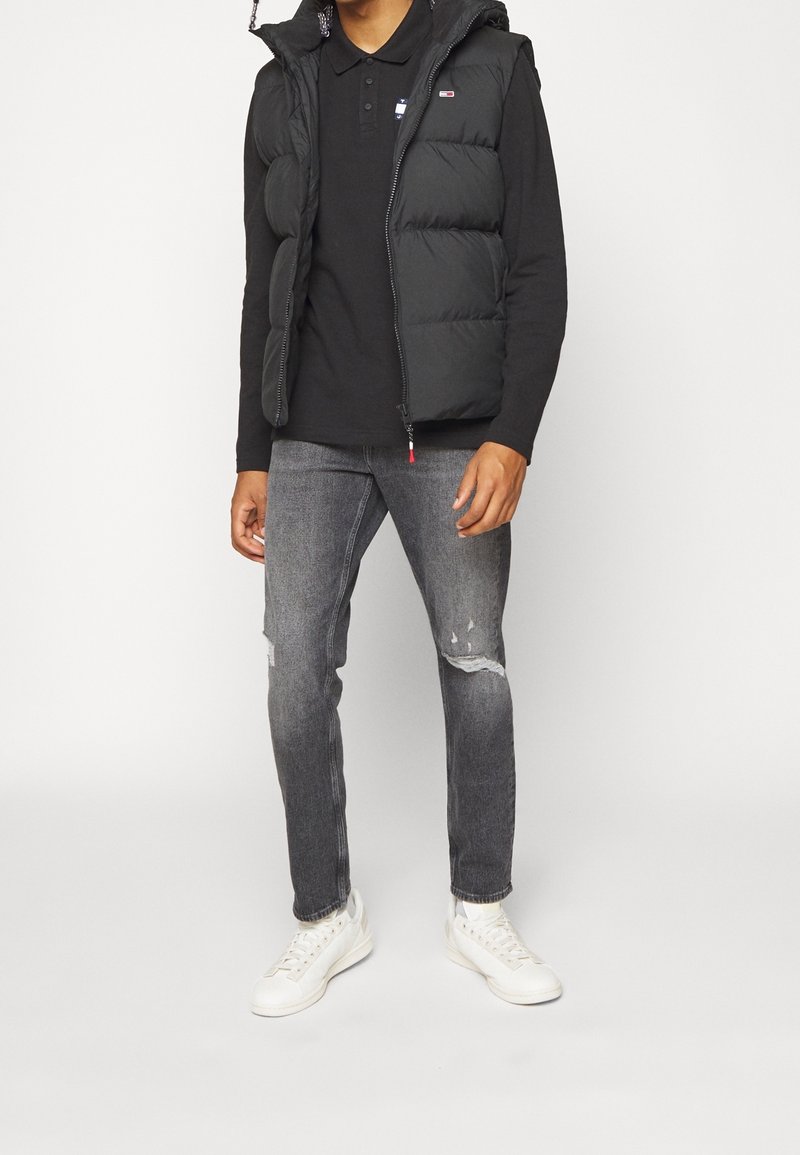 Svart pufferväst med blixtlås i kragen, matchad med svart långärmad pikétröja, grå slitna jeans och vita sneakers.