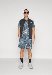 Tenue de sport comprenant un gilet sans manches noir, une chemise à motifs verts et bleus, un short assorti, des chaussettes noires et des baskets légères.
