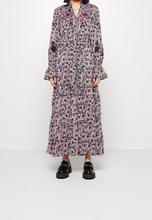 Kobieta w długiej sukience w kwiatowy wzór z falbankami na rękawach i brzegu, w czarnych masywnych mokasynach, stoi na tle białego tła.