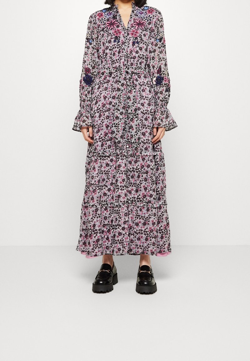 Femme portant une longue robe à motif floral avec des manches et un ourlet à volants, associée à des mocassins noirs épais, debout devant un fond blanc.